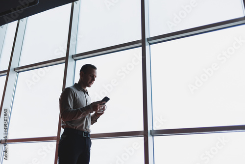 A young executive is holding a phone while standing in an office interior and looking out a large window overlooking the city. Male manager with a phone is thinking about a new business project