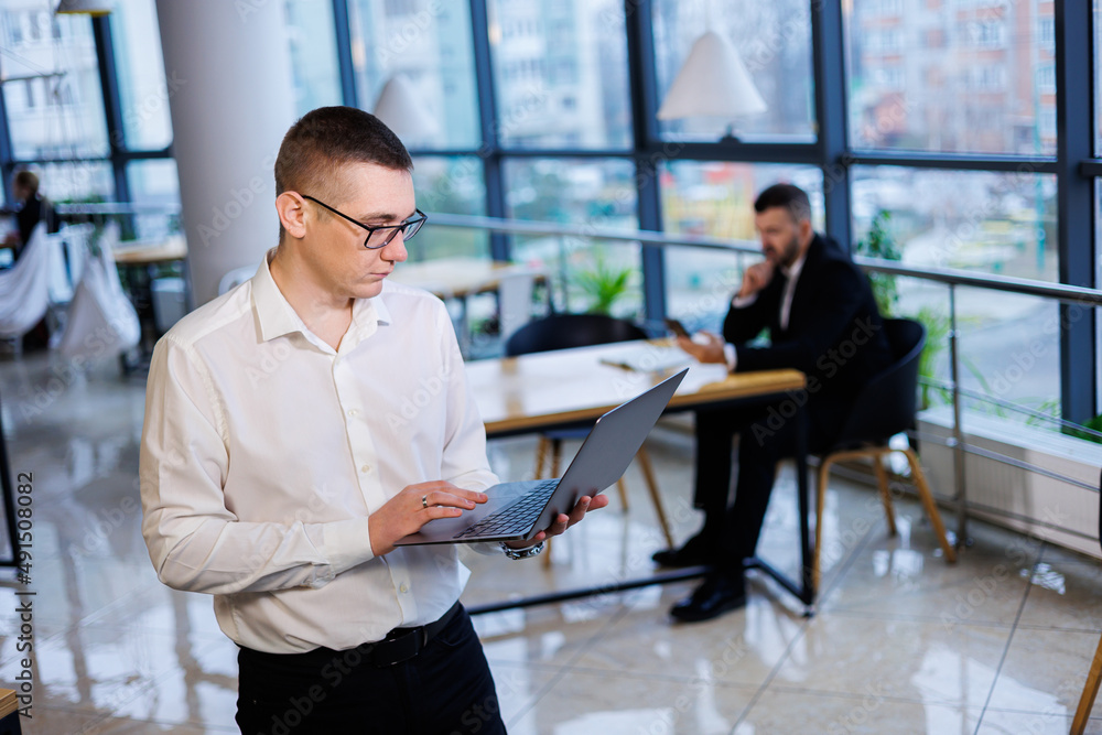 A young businessman man in glasses and a white shirt with a laptop in his hands stands in the office and works