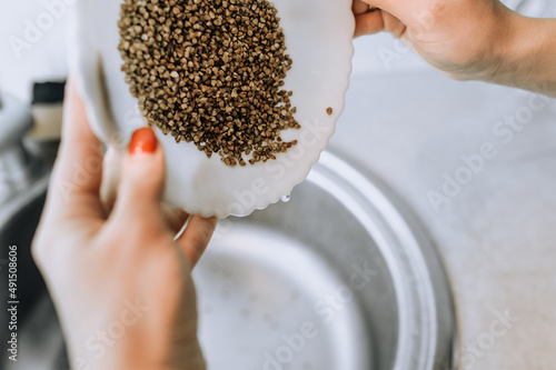 The girl, the cook, holds a plate with grain in her hands and rinses buckwheat with water over the washbasin. Cooking in the kitchen. photo