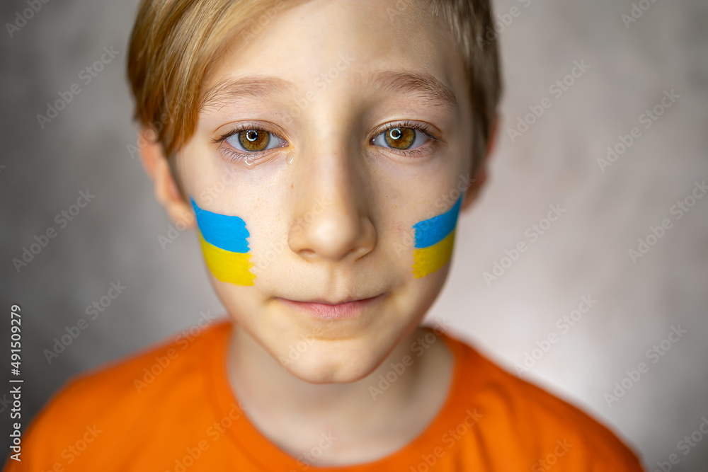 a sad child against the war, a boy with tears in his eyes and a painted flag of Ukraine on his cheeks looks at the camera with pity