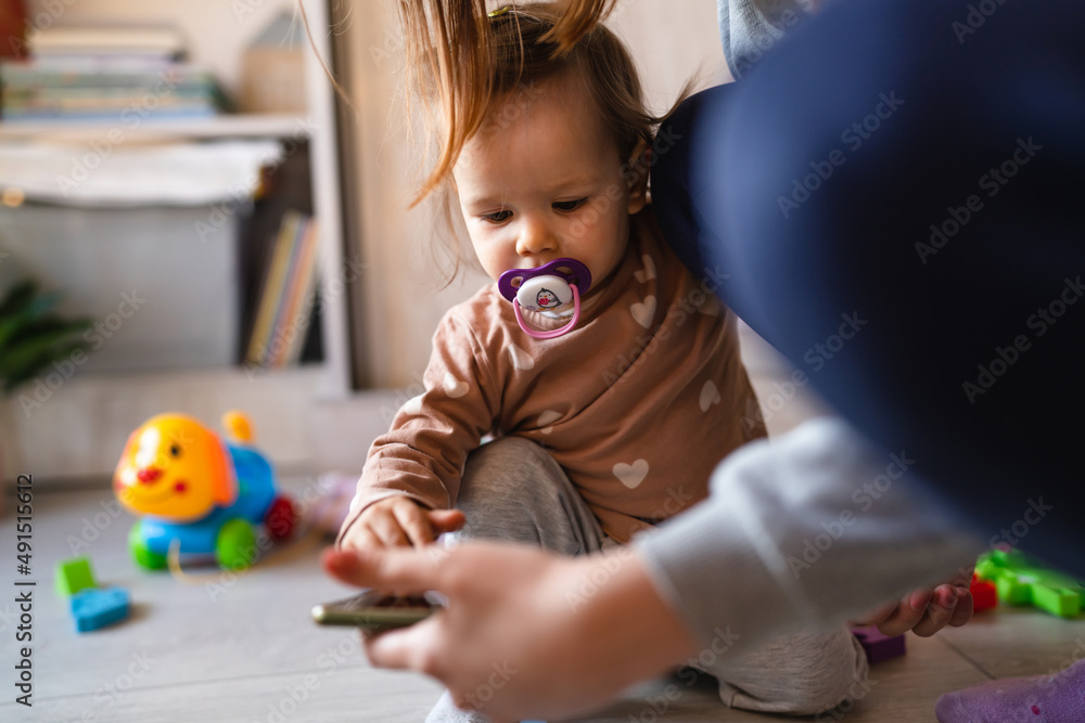 One baby small caucasian infant girl playing on the floor at home copy space holding mobile phone smartphone using to call or watch video in day front view One baby small caucasian infant girl playing on the floor at home copy space holding mobile phone smartphone using to call or watch video in day front view