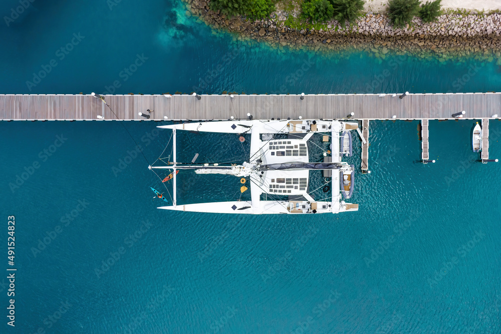 Luxurious white modern expensive catamaran yacht moored to a long pier in a bay with crystal clear turquoise ocean water. Aerial view. Tourism concept