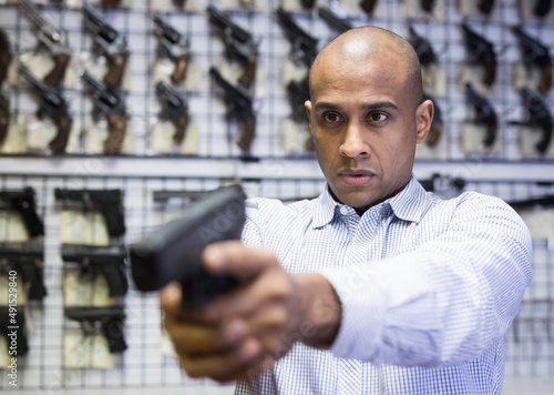 Focused latin american man examining convenience and accuracy of new pistol in gun shop, imitating aiming