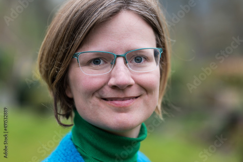 Close up portrait of a young white woman with glasses wearing a blue and green sweater with a green blurry nature background