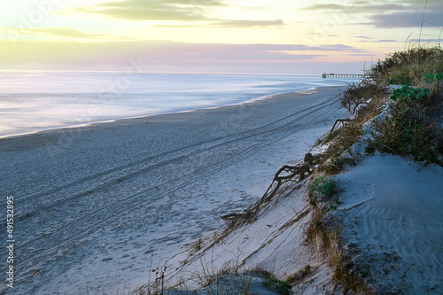 Sunset at the dunes and beach with a distant pier over a smooth ocean with orange, yellow, and pink sky