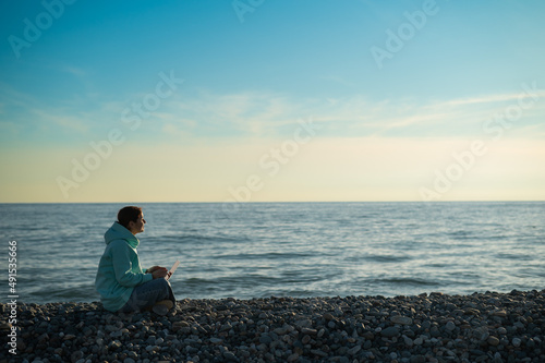 Happy caucasian woman working on a laptop while sitting on a pebble beach.