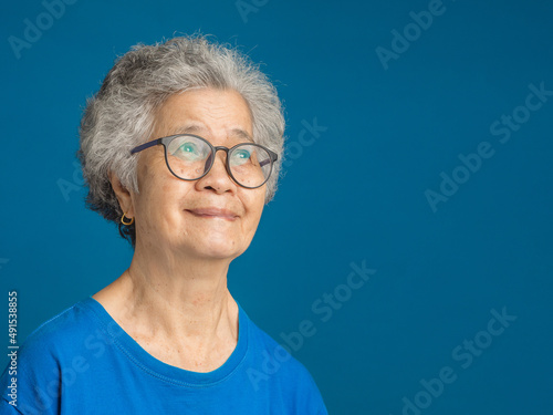 Senior woman with short gray hair wearing glasses and looking up with a smile while standing on a blue background