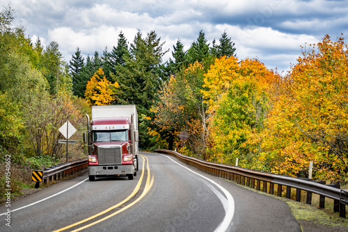 Dark red classic powerful big rig semi truck transporting commercial cargo driving on the winding road through the autumn forest