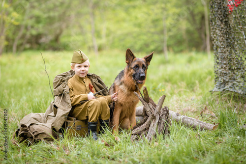 a boy in a military uniform in a clearing  sitting by a campfire with a German shepherd.Two friends defend the motherland