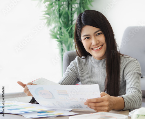 Businesswoman sitting in office busy with graph report analysis looking at camera smiling, portrait. Asian modern executive working woman review corporate finance and marketing paper chart document.