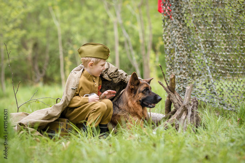 a boy in a military uniform in a clearing  sitting by a campfire with a German shepherd.Two friends defend the motherland
