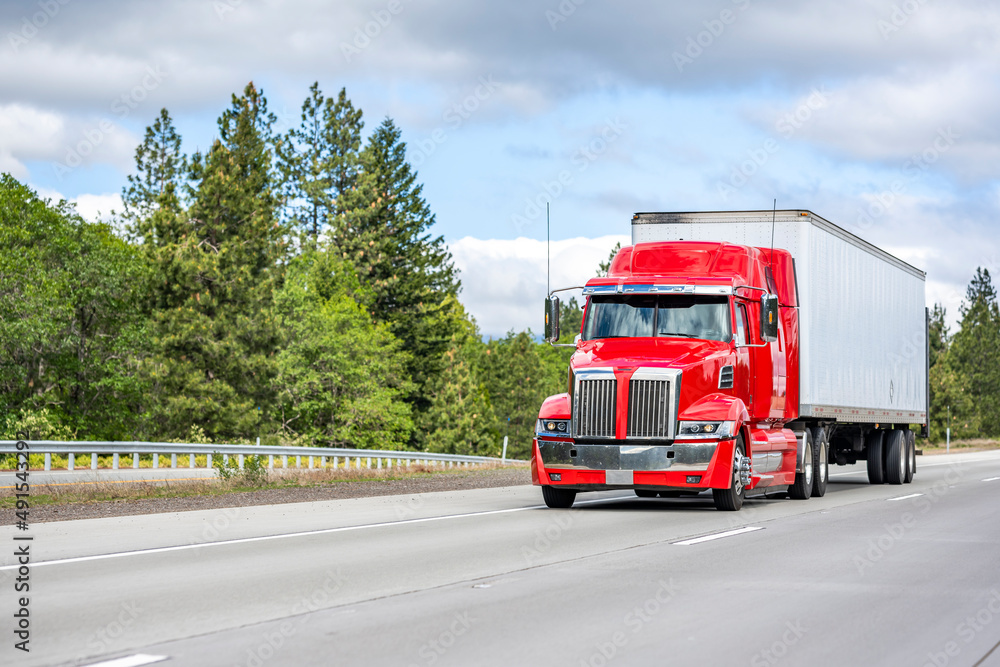 Stylish bright red big rig semi truck with chrome parts transporting cargo in dry van semi trailer running on the straight wide highway road with green trees on the side