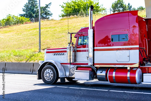 Shiny red big rig classic bonnet semi truck tractor with long truck driver rest compartment and chrome parts transporting cargo running on the sunny highway road