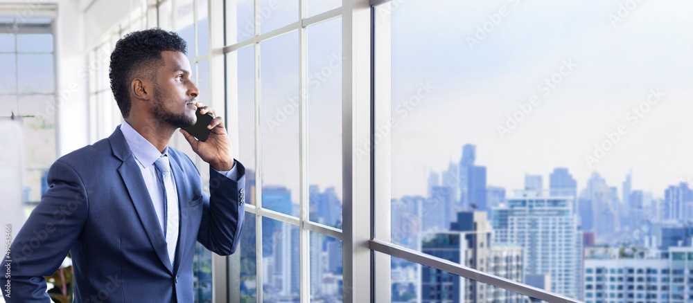 African American businessman in formal suit using mobile phone while looking out the window to the cityscape with skyscraper view for vision, dream and successful business and investment planning African American businessman in formal suit using mobile phone while looking out the window to the cityscape with skyscraper view for vision, dream and successful business and investment planning