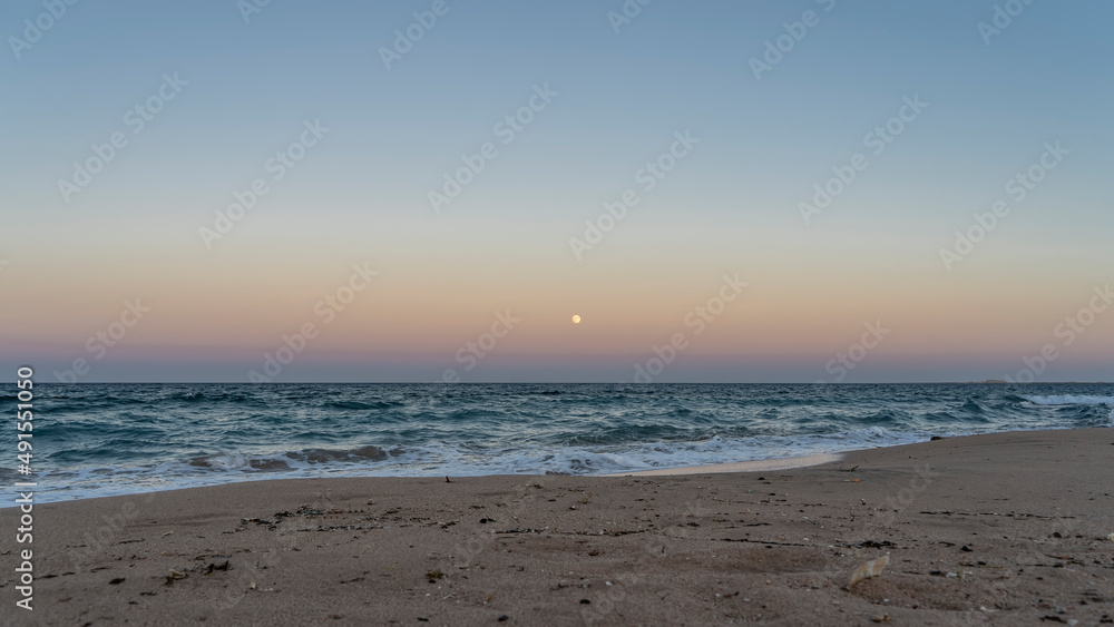 Evening on the red sea beach. The full moon is shining in the pinkish-blue sky. Turquoise waves roll onto the sandy shore and foam. Egypt.