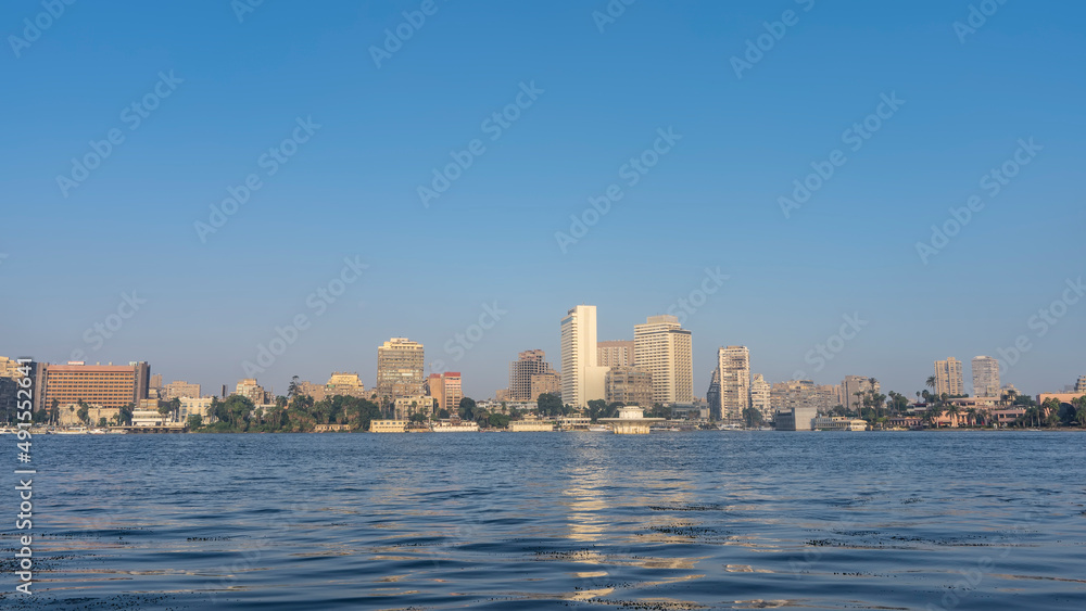 On the banks of the Nile in Cairo, city houses and green vegetation are visible. Ripples and reflections on the surface of blue water. Clear azure sky. Egypt.