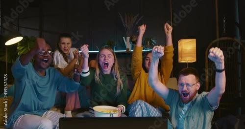 Happy young multiethnic sports fans sit together watching football on TV at home with snacks celebrating goal and win.