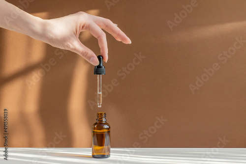 Pipette with cosmetic serum in women's hands on light brown background, in rays of sunlight. Concept of body care, rejuvenation. Close-up, copy space
