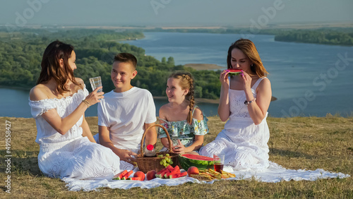 Mother and happy children have festive picnic with tasty food sitting on white tablecloth on green brae against tranquil wide river on sunny summer day