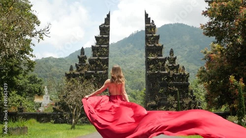 Pretty asian woman walks to balinese gates in long red dress in slowmotion on Hangara gates in Bali Indonesia. High quality fullhd footage
