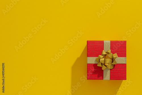 One red gift box with gold ribbon and bow on yellow table background. Minimal festive gift background. Top view. Flat lay.	
