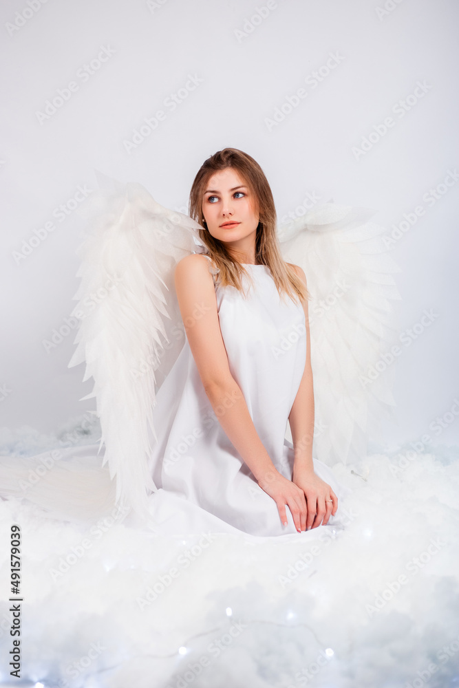 An attractive girl in a white tunic with large white wings behind her back poses while sitting in white luminous clouds.
An attractive girl in a white tunic with large white wings behind her back poses while sitting in white luminous clouds.