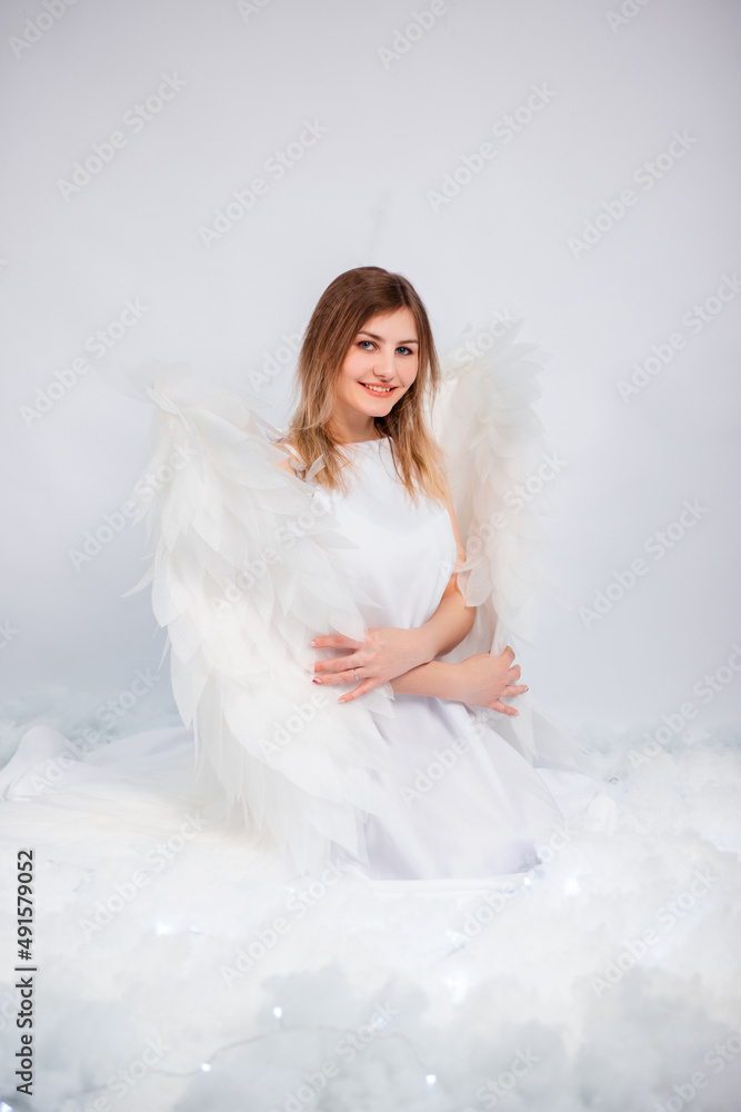 An attractive girl in a white tunic with large white wings behind her back poses while sitting in white luminous clouds.
An attractive girl in a white tunic with large white wings behind her back poses while sitting in white luminous clouds.