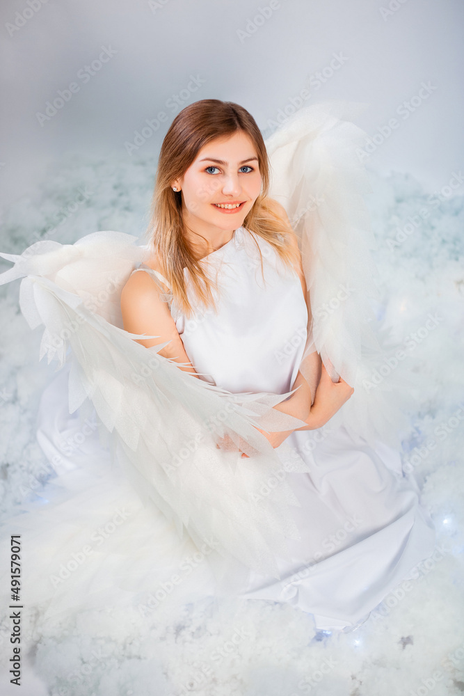 An attractive girl in a white tunic with large white wings behind her back poses while sitting in white luminous clouds.
An attractive girl in a white tunic with large white wings behind her back poses while sitting in white luminous clouds.