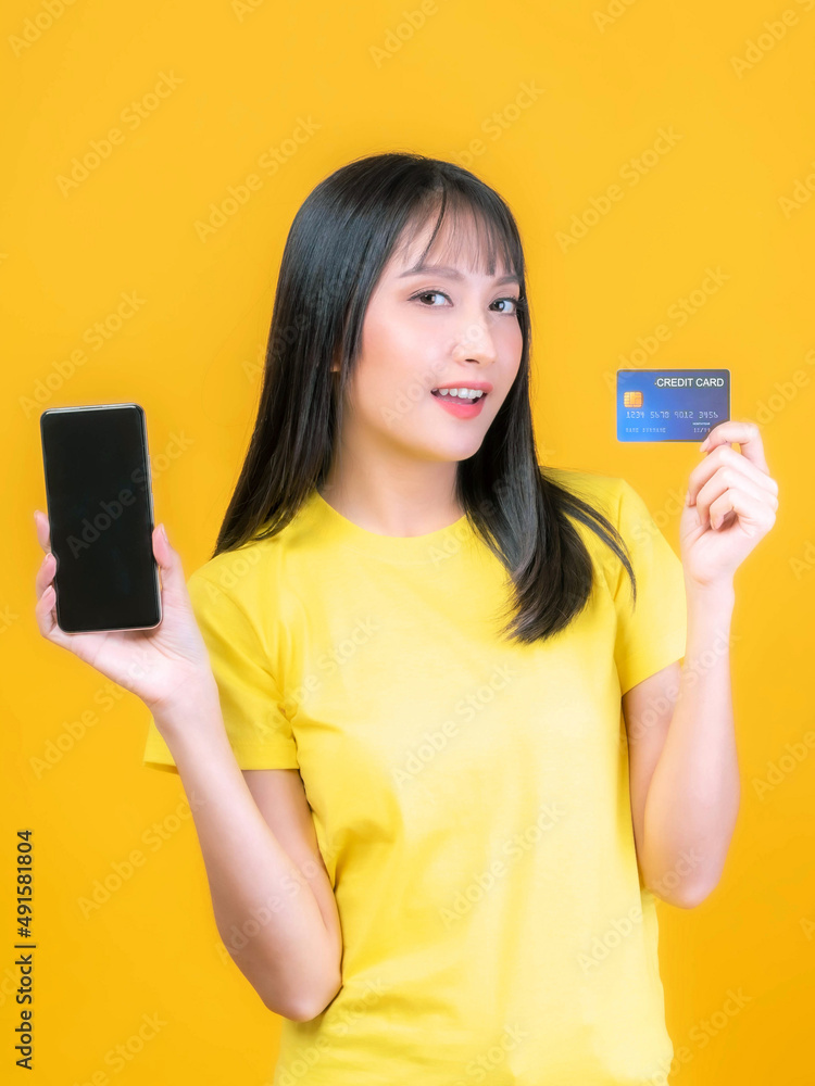 beautiful Asian young woman cute girl with bangs hair style in yellow shirt holding credit card and smartphone ,mobile phone uses a online banking for pay online shopping isolated on yellow background beautiful Asian young woman cute girl with bangs hair style in yellow shirt holding credit card and smartphone ,mobile phone uses a online banking for pay online shopping isolated on yellow background