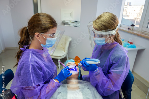 the dentist with his assistant in overalls apply a UV lamp to the patient's teeth.