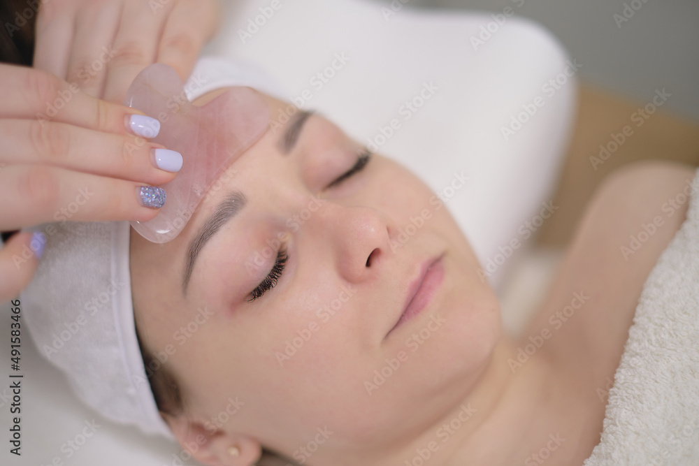 Calm patient woman undergoing the cosmetic facial massage procedures. Woman in salon making beauty treatment with gua sha stone Calm patient woman undergoing the cosmetic facial massage procedures. Woman in salon making beauty treatment with gua sha stone