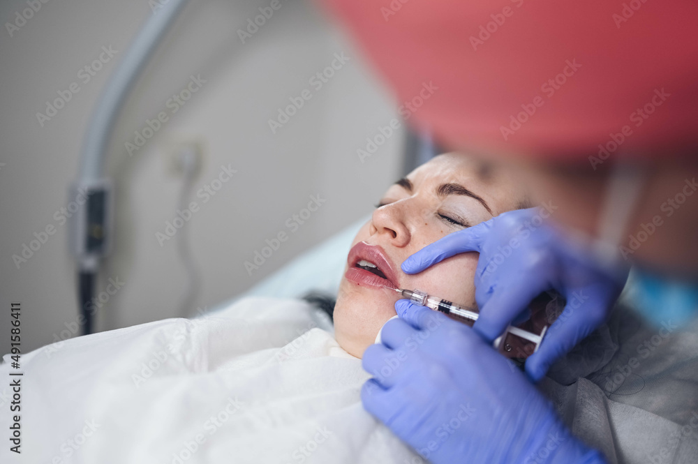 Cosmetologist doing painful lip augmentation procedure with hyaluronic acid. The beautician pierces lips by needle. Woman suffering subcutaneous injection to increase lips shape with dermal filler. Cosmetologist doing painful lip augmentation procedure with hyaluronic acid. The beautician pierces lips by needle. Woman suffering subcutaneous injection to increase lips shape with dermal filler.