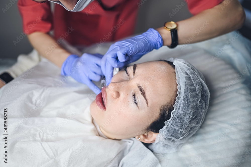 Cosmetologist doing painful lip augmentation procedure with hyaluronic acid. The beautician