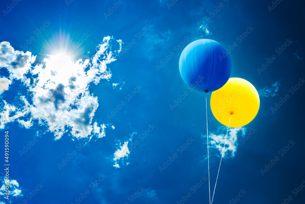 Two balloons in ukrainian flag colors over blue sky and sun from behind the clouds.