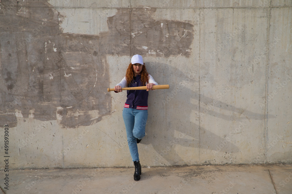 young and beautiful redhead woman is happy with baseball cap, jacket, baseball bat and jeans, she is posing in front of grey concrete background. Concept sport and recreations.