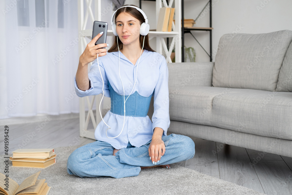 Video call. Smiling woman. Online education. Pretty happy casual lady in headphones looking mobile phone in hand sitting floor light room interior.