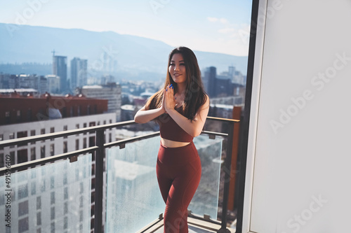 Beautiful Asian smiling fit woman doing yoga on sunny balcony in sport suit. Young happy lady on terrace at summertime. Big city view. Sports exercises and gymnastics, healthy way of life concept
