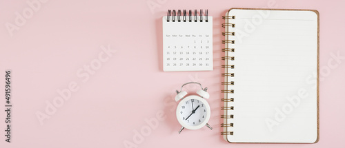 top view of blank opened note book, g white monthly calendar and analog alarm clock on sweet pink background