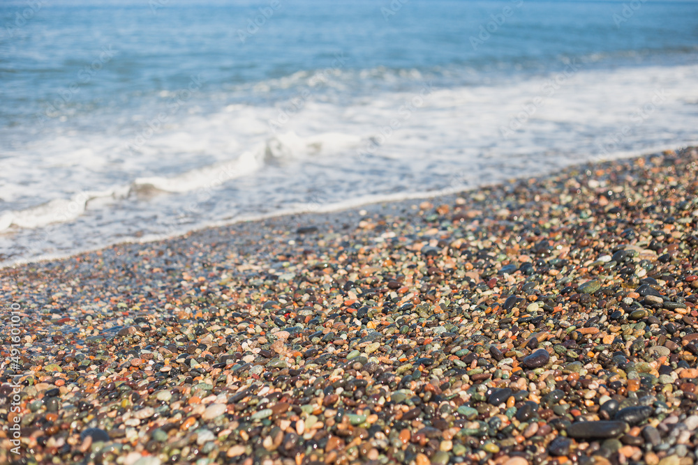 Diagonal coastline of the sea with a pebble beach - colored pebble stones wet from the wave in the foreground and blue water Diagonal coastline of the sea with a pebble beach - colored pebble stones wet from the wave in the foreground and blue water