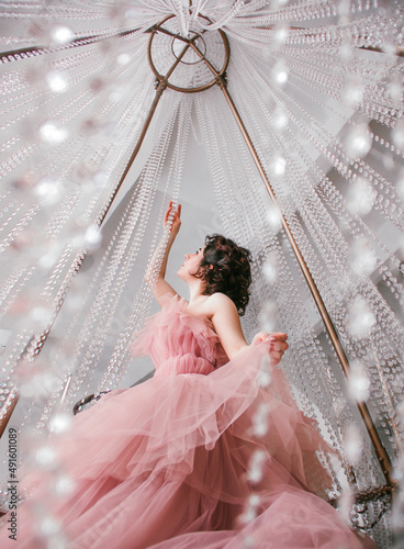 young woman with short black hair in pink princess dress is sitting on the crystal chandelier and looking up on the white ceiling background, bottom camera. fashion concept, free space