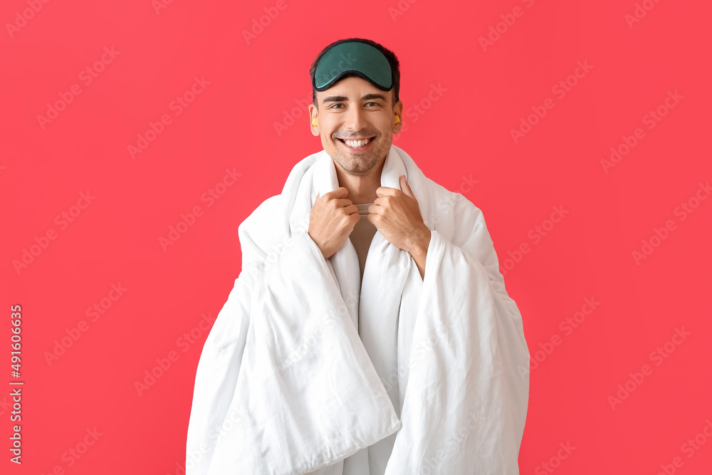 Smiling young man with sleeping mask, blanket and ear plugs on red background Smiling young man with sleeping mask, blanket and ear plugs on red background