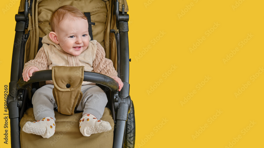 A happy child is sitting in a baby carriage on a studio yellow background. Smiling toddler baby boy in a transformer stroller for transporting children, age eight months A happy child is sitting in a baby carriage on a studio yellow background. Smiling toddler baby boy in a transformer stroller for transporting children, age eight months