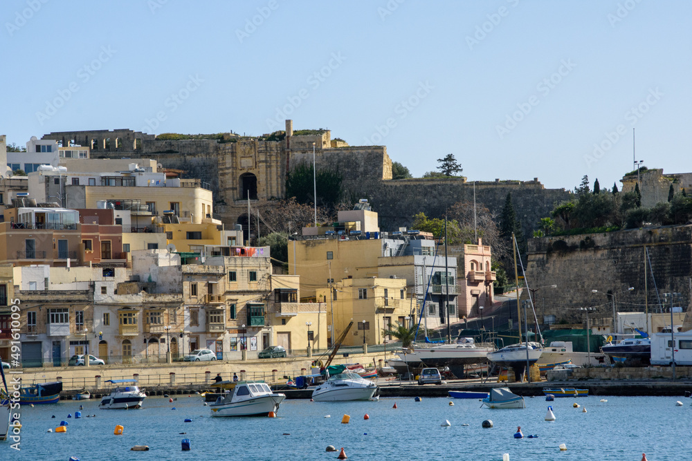Kalkara village in Malta, with boats moored on Kalkara Creek. In the background are parts of the  Cottonera Lines fortifications including the Our Saviour Gate and its small derelict Chapel.