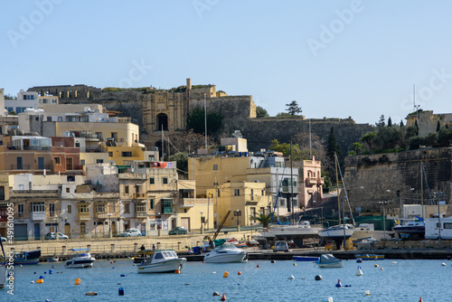 Fototapeta Naklejka Na Ścianę i Meble -  Kalkara village in Malta, with boats moored on Kalkara Creek. In the background are parts of the  Cottonera Lines fortifications including the Our Saviour Gate and its small derelict Chapel.