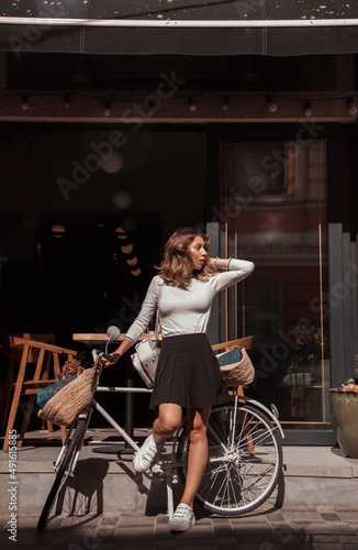 young curly brunette girl in white sweater and black skirt is standing in modern cafe on terrace near bicycle and looking away with hand near hair on showcase background. lifestyle concept, free space