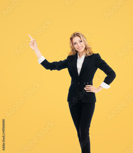 Full body of smiling executive woman in black confident suit, pointing showing copyspace. Business concept. Yellow background. Young cheerful businesswoman.