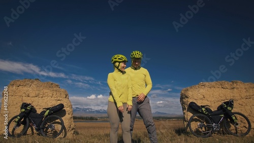 The man and woman travel on mixed terrain cycle bike touring with bikepacking. The two people journey with bicycle bags. Sport sportswear in green black colors. Mountain snow capped, stone arch. photo