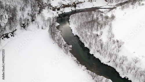 Beautiful clear river with meltwater flowing gently between the thin trees covered in a thick layer of snow Hokkaido Province in Japan. High angle drone dolley shot photo