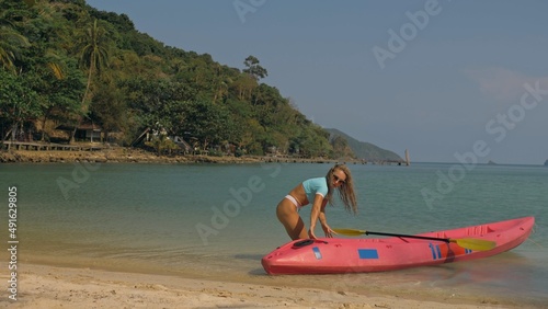 Slim young woman pushes pink plastic canoe to clear ocean water from sandy beach at exotic resort on sunny day. Traveling to tropical countries. Attractive sexy girl posing near kayak.