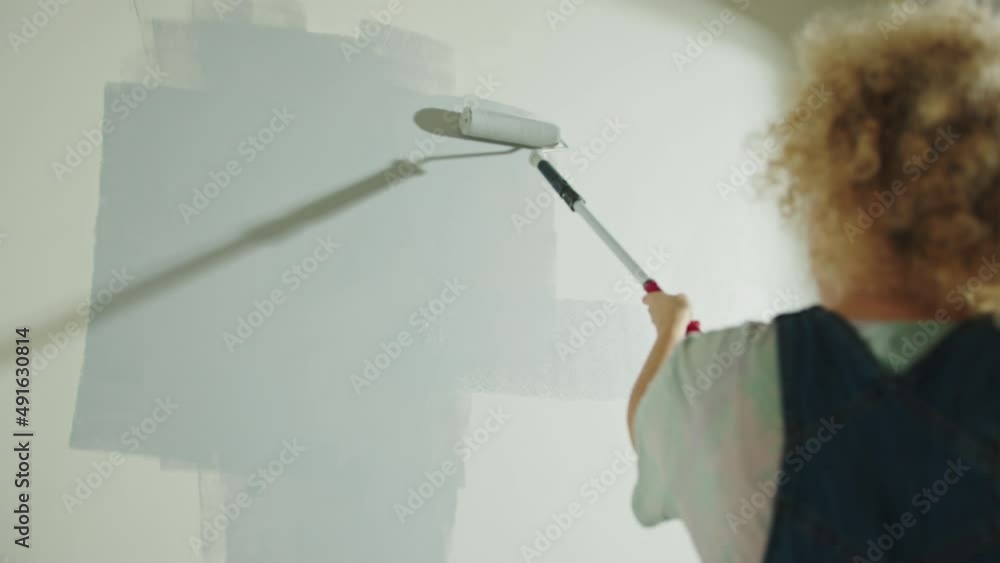 Curly hair Woman is painting wall in gray using paint rollers. View from behind Curly hair Woman is painting wall in gray using paint rollers. View from behind